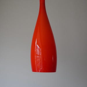 Puede incluir: Una lámpara colgante de vidrio naranja brillante que cuelga de un cable blanco. La lámpara tiene forma de lágrima.
