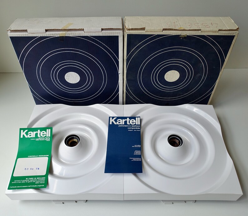 Puede incluir: Dos l&aacute;mparas de techo blancas Kartell con dise&ntilde;os circulares y una caja azul y blanca con el logotipo de Kartell. Las cajas tienen un dise&ntilde;o de c&iacute;rculos conc&eacute;ntricos en azul y blanco.