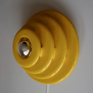 May include: A yellow wall sconce with a layered, circular design. The sconce has a glossy finish and a silver-colored, spherical light fixture in the center. A white cord extends from the bottom.
