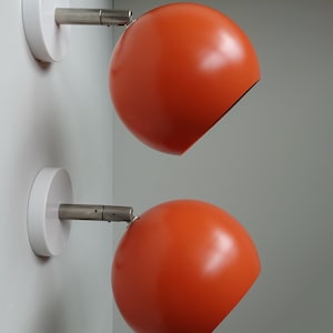 Puede incluir: Dos apliques de pared de color naranja con bases circulares blancas y brazos ajustables plateados. Los apliques están montados en una pared blanca.