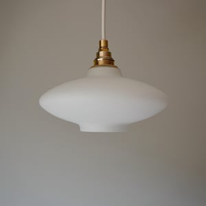May include: A pendant light fixture featuring a wide, saucer-shaped frosted white glass shade. The lamp is suspended by a white cord and includes a metallic brass-toned socket assembly at the top where the shade connects to the electrical cord.