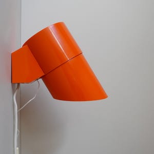 Peut inclure: Une lampe murale de couleur orange vif avec un design cylindrique. La lampe est inclinée vers le bas et possède un cordon blanc.