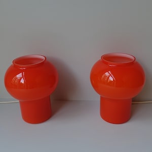 May include: Two orange glass table lamps with white cords and switches. The lamps have a round, bulbous top and a cylindrical base.