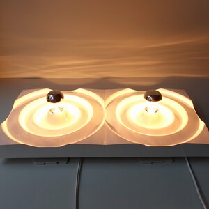 Puede incluir: Una l&aacute;mpara de pared blanca moderna con dos luminarias circulares. Cada luminaria tiene una c&uacute;pula cromada y un difusor circular blanco texturizado. Las luces est&aacute;n encendidas y emiten un brillo amarillo c&aacute;lido.