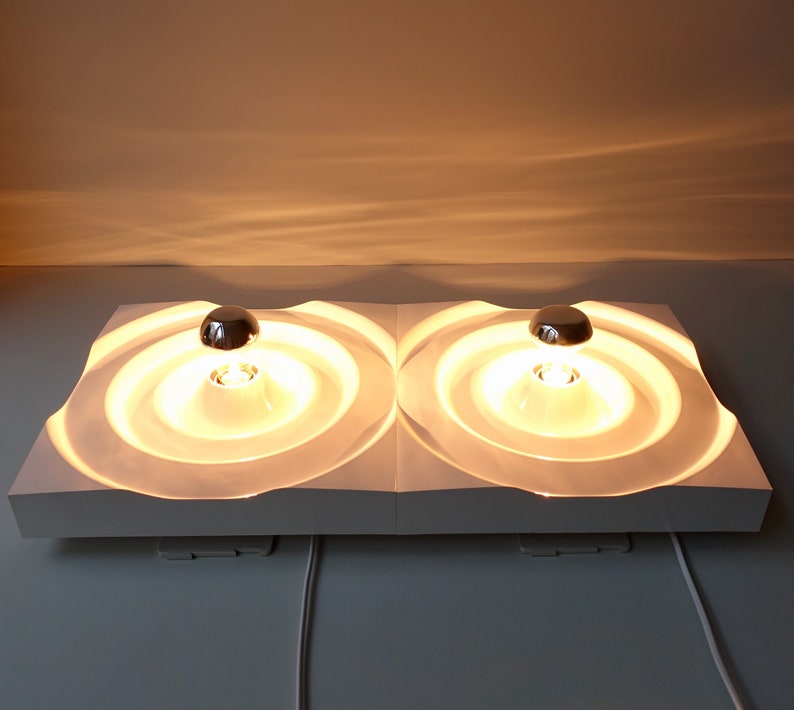 Puede incluir: Una l&aacute;mpara de pared blanca moderna con dos luminarias circulares. Cada luminaria tiene una c&uacute;pula cromada y un difusor circular blanco texturizado. Las luces est&aacute;n encendidas y emiten un brillo amarillo c&aacute;lido.