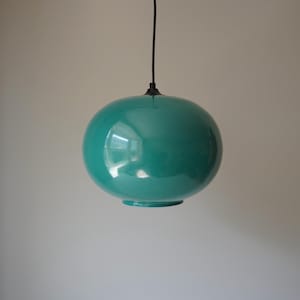 Puede incluir: Una lámpara colgante esférica de color turquesa. La pantalla brillante y redondeada cuelga de un cable negro. La luz refleja el entorno. La lámpara tiene un diseño sencillo y moderno, adecuado para varios estilos de interior.