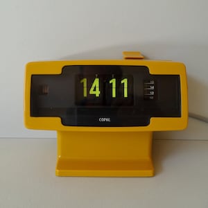 May include: A yellow retro flip clock displaying the time 14:11 in green digits. The clock has a rectangular face with a black display area and the brand name "COPAL" printed below the time. The clock is made of plastic.