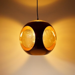 May include: A modern pendant light fixture with a spherical design. The light has a dark brown exterior with two large, golden-colored, oval-shaped openings. The top and interior of the openings have a gold leaf finish. The light hangs from a black cord.