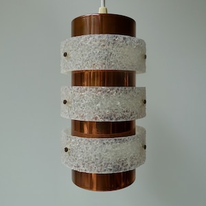 Könnte beinhalten: Eine kupferne und weiße Pendelleuchte mit einem strukturierten Glasschirm. Die Leuchte hat drei Glasstufen und ist an einem weißen Kabel aufgehängt.