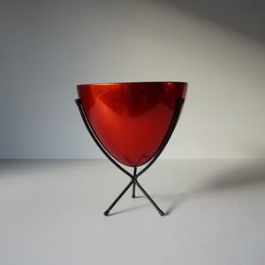 May include: A red metal planter with a wide, rounded base and a narrow top, sitting on a black metal stand with three legs.