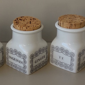 Puede incluir: Conjunto de ocho tarros de especias de cerámica blanca con tapas de corcho. Cada tarro tiene un borde decorativo blanco y negro y una etiqueta con el nombre de la especia: clavo, té, jengibre, canela, alcaravea, lager blad.