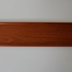 Puede incluir: Bandeja rectangular de madera en un tono marrón rojizo intenso. Presenta una zona central rebajada y extremos angulados. La veta de la madera es visible, realzando su aspecto natural. Mide aproximadamente 50 cm de largo.