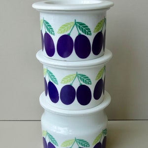 Può includere: Un set di tre barattoli di stoccaggio in ceramica bianca con un design a prugne. I barattoli sono impilati uno sopra l'altro. Ogni barattolo ha un coperchio bianco e un design di prugne viola con foglie verdi.