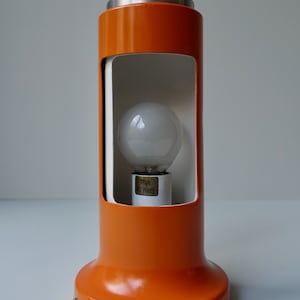 Puede incluir: Lámpara de mesa naranja y blanca con base de metal y cable blanco. La lámpara tiene una pantalla de vidrio transparente y una bombilla en el interior.