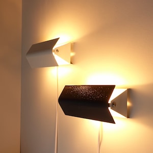 May include: Two wall-mounted lights, one white and one black, with a simple, modern design. The lights are turned on and illuminating the wall.
