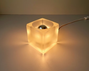 May include: A frosted glass cube lamp with a silver light bulb inside. The lamp is lit and casting a warm glow. The lamp has a white cord with a switch.