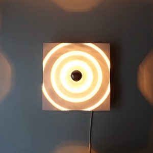 Puede incluir: Una l&aacute;mpara de pared circular blanca con anillos conc&eacute;ntricos y una sola bombilla en el centro.