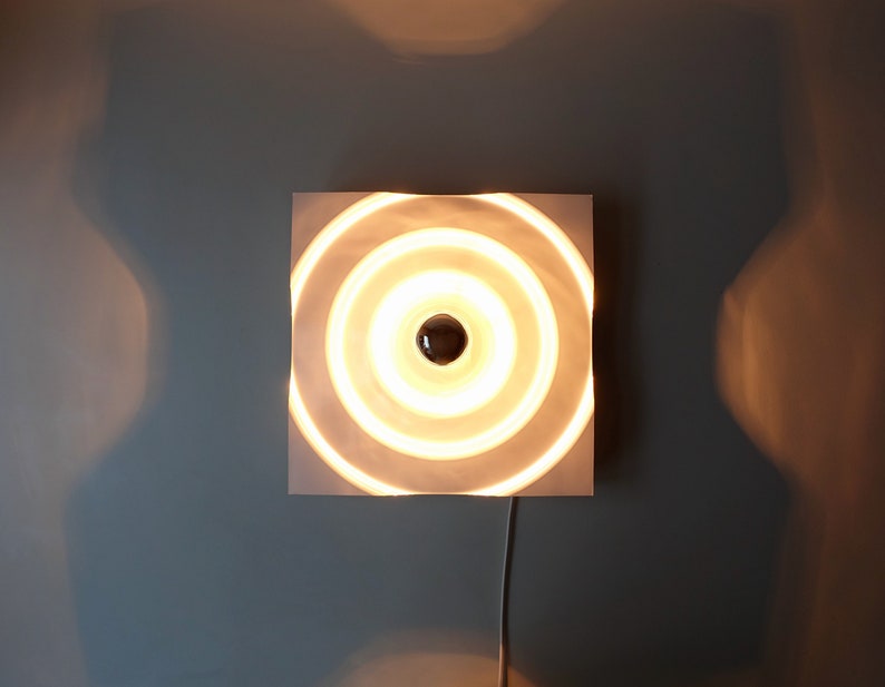 Puede incluir: Una l&aacute;mpara de pared circular blanca con anillos conc&eacute;ntricos y una sola bombilla en el centro.