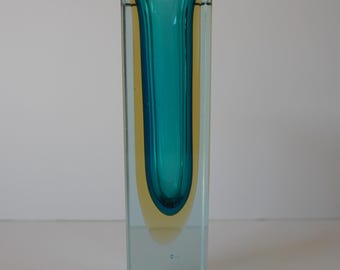 May include: A rectangular glass vase with a teal interior and a yellow-toned exterior. The vase is transparent, allowing light to pass through, and has a simple, modern design. The vase is approximately 8 inches tall.