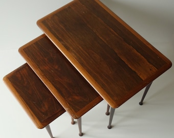 Puede incluir: Un conjunto de tres mesas nido de madera oscura con un borde de madera más clara. Las mesas tienen forma rectangular y están sostenidas por cuatro patas delgadas.