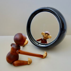 Op de afbeelding: Een houten aapfiguur dat in een ronde zwarte spiegel kijkt. De aap is bruin met een lichtbruin gezicht en heeft een lichtbruine houten neus. De spiegel is zwart met een zilveren reflecterend oppervlak.