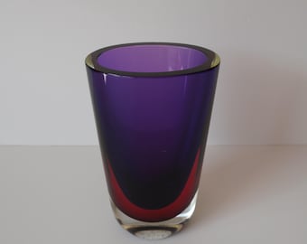 May include: A tall, clear glass vase with a vibrant purple interior and a red curved accent at the base. The rim has a thin, yellow-green band. The vase is set against a plain white background.