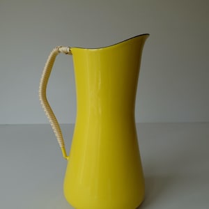 Puede incluir: Una jarra de esmalte amarillo con un asa tejida. La jarra tiene una boca ancha y una base redondeada.