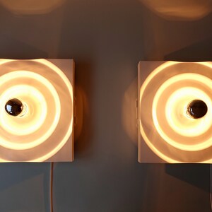 Puede incluir: Dos apliques de pared blancos con un dise&ntilde;o circular de anillos conc&eacute;ntricos. Cada aplique tiene una sola bombilla en el centro.