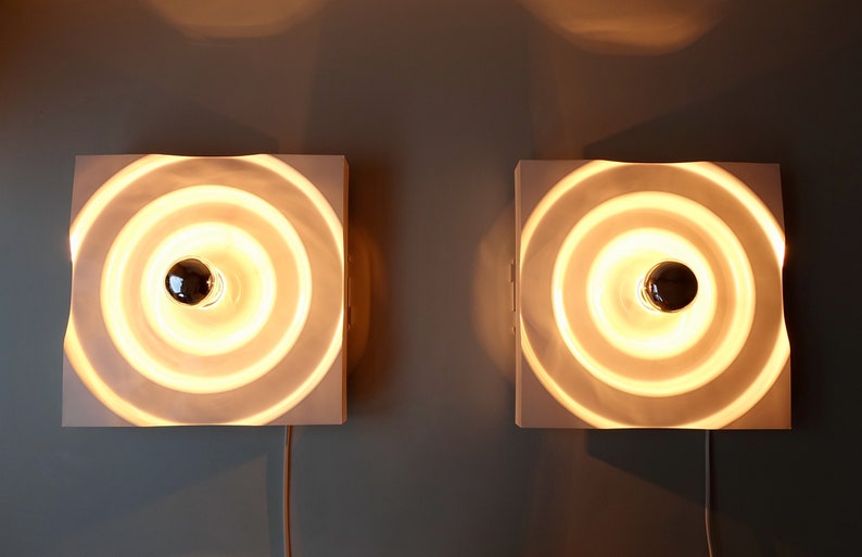 Puede incluir: Dos apliques de pared blancos con un dise&ntilde;o circular de anillos conc&eacute;ntricos. Cada aplique tiene una sola bombilla en el centro.