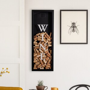 May include: Black framed wall decor with the word "WINE" spelled out using cork stoppers. The decor is hanging on a white wall above a dining table with chairs.