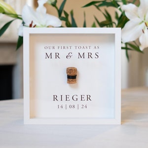May include: White frame with a champagne cork inside. The frame has the text "Our First Toast as Mr & Mrs Rieger 14 | 08 | 24" printed on it.