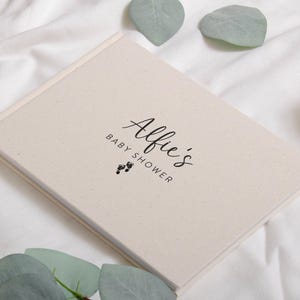 May include: A cream-colored baby shower guest book with the name "Alfie's" in elegant script, along with the words "Baby Shower" and tiny footprint graphics. The book is rectangular and lies on a white surface, with eucalyptus leaves.