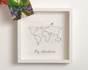 May include: White picture frame with a world map drawn in one line with a small airplane flying over it. The text "My Adventures" is written below the map.