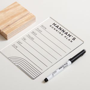 May include: A clear acrylic running plan board with the text "HANNAH'S RUNNING PLAN" and days of the week. Includes a black dry-erase marker and a wooden stand. The board has a curved line design at the bottom.