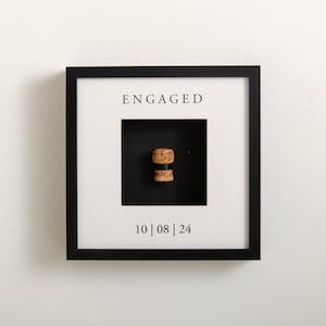 Puede incluir: Una imagen enmarcada en negro con un paspartú blanco y un corcho de una botella de champán en su interior. La imagen dice "ENGAGED" y "10|08|24" en texto negro.
