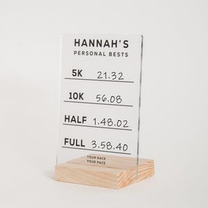 Könnte beinhalten: Ein klares Acrylschild mit schwarzem Text, der "HANNAH'S PERSONAL BESTS" lautet und Laufzeiten für 5K, 10K, Halbmarathon und Marathon auflistet. Das Schild steht auf einem Holzsockel mit dem Text "YOUR RACE YOUR PACE".