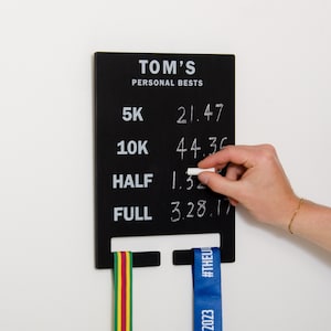 May include: Black chalkboard with the text "TOM'S PERSONAL BESTS" and a list of running distances with times written in white chalk. The distances are 5K, 10K, HALF, and FULL. A hand is holding a piece of chalk and writing on the board. Two medal ribbons are hanging on hooks below the board.