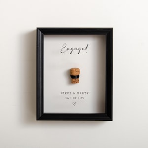 Può includere: Un ricordo di fidanzamento incorniciato con un tappo di champagne. La parola "Engaged" è scritta in corsivo in alto. Sotto il tappo ci sono i nomi "Nikki & Marty", la data e un simbolo a forma di cuore. La cornice è nera.