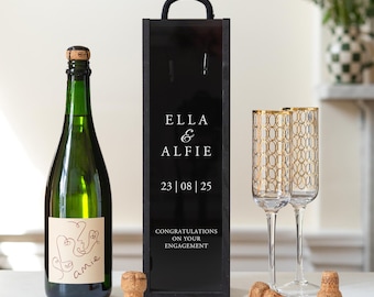 May include: A green glass bottle with a beige label featuring a line-art face, a tall black wooden box with a rope handle printed with ELLA & ALFIE 23 | 08 | 25 CONGRATULATIONS ON YOUR ENGAGEMENT, two gold-patterned champagne flutes, and three corks.