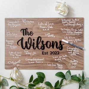Könnte beinhalten: Ein Holzschild mit den Worten "The Wilsons Est 2022" in schwarzer Schrift. Das Schild ist mit weißen handgeschriebenen Botschaften von Familie und Freunden bedeckt, darunter "Herzlichen Glückwunsch!" und "Wir lieben euch beide!"