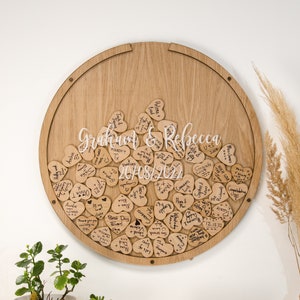 May include: A circular wooden board with a groove around the edge, holding a collection of small wooden hearts. Each heart has a handwritten message on it, including the names "Graham & Rebecca" and the date "20/08/2022".