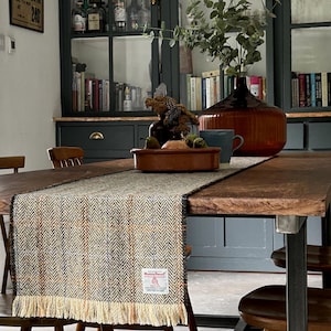 May include: A wooden dining table with a brown tweed table runner. The table has a dark wood top and black metal legs. There are four chairs around the table, two of which are visible in the image. The table is set with a bowl of fruit and a vase of greenery.