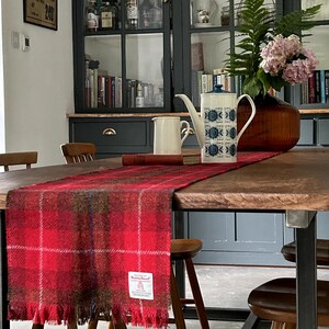 Peut inclure: Un chemin de table en tartan rouge et vert avec des franges sur les bords est posé sur une table en bois avec des pieds en métal noir. Le chemin de table porte une étiquette blanche avec le texte "Harris Tweed". La table est dressée avec deux chaises et un chemin de table en bois.