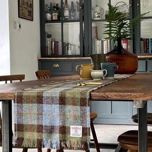 Puede incluir: Una mesa de comedor rústica de madera con un camino de mesa a cuadros en tonos marrones, azules y verdes. La mesa está decorada con tazas, una jarra y un jarrón decorativo con un helecho. Sillas de madera rodean la mesa y un armario verde oscuro está en el fondo.