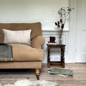 May include: A tan linen sofa with a beige throw pillow and a dark green throw blanket. The sofa is in a living room with a wooden side table, a vase of dried flowers, and a book on the floor.