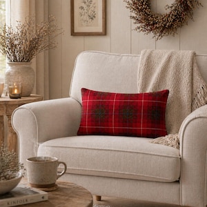 Op de afbeelding: Een rood geruit lendenkussen met een donkergroen en bruin bloemmotief ligt op een crèmekleurige fauteuil. Een beige deken met franjes is over de stoel gedrapeerd. Een koffiemok en een boek liggen op een bijzettafel.