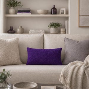 Op de afbeelding: Een moderne woonkamer met een beige bank met decoratieve kussens. Een levendig paars rechthoekig kussen is het middelpunt. Andere kussens zijn beige en grijs. Een beige plaid met franjes is over de bank gedrapeerd.