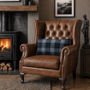 Op de afbeelding: Een bruine leren wingback fauteuil met een gecapitonneerde rugleuning en een geruit lendenkussen. De stoel heeft donkere houten poten en staat voor een open haard. Het kussen heeft een blauw en grijs ruitpatroon.