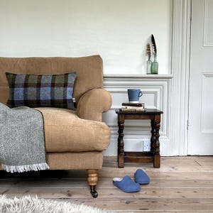 May include: A tan coloured sofa with a plaid throw pillow and a grey throw blanket. A small wooden side table with a blue mug and books sits next to the sofa. Two blue slippers are on the wooden floor.