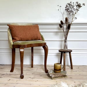 May include: A vintage wooden chair with a green velvet seat and an orange cushion. The chair has four legs and a curved back. The chair is sitting on a wooden floor with a small wooden stool and a stack of books in front of it.
